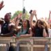 Crowd of festivalgoers standing behind a metal safety barrier at an outdoor music event, many with their hands raised in the air. A group at the front includes several young adults leaning on the barrier, some holding drinks and mobile phones, with trees and stage equipment visible in the background under bright daylight.