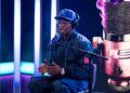 Cam'ron wearing a "KC" cap and headphones speaks into a microphone on a podcast set with vibrant lighting and a large microphone graphic.
