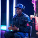 Cam'ron wearing a "KC" cap and headphones speaks into a microphone on a podcast set with vibrant lighting and a large microphone graphic.