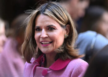A woman with shoulder-length hair wearing a pink coat is smiling in a crowd.