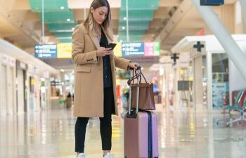 Woman looking at her smartphone to find her flight information while navigating an airport terminal with her luggage in tow.