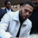Chris Brown, in a textured white jacket, signing an autograph, surrounded by people in formal attire outdoors.