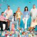 An image of a group of individuals, both standing and seated, gathered around a colorful display of various objects and confetti against a vibrant blue backdrop.