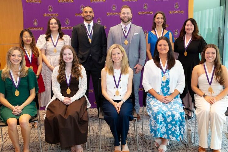 Group of award recipients wearing medals pose for a photo in front of a University of Scranton backdrop.