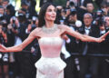 Demi Moore in a strapless shimmering gown poses with arms outstretched on a red carpet while photographers crowd behind her taking pictures.