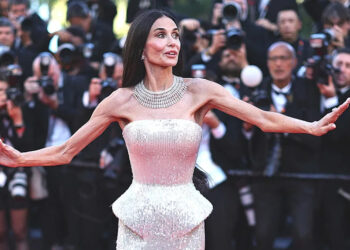 Demi Moore in a strapless shimmering gown poses with arms outstretched on a red carpet while photographers crowd behind her taking pictures.