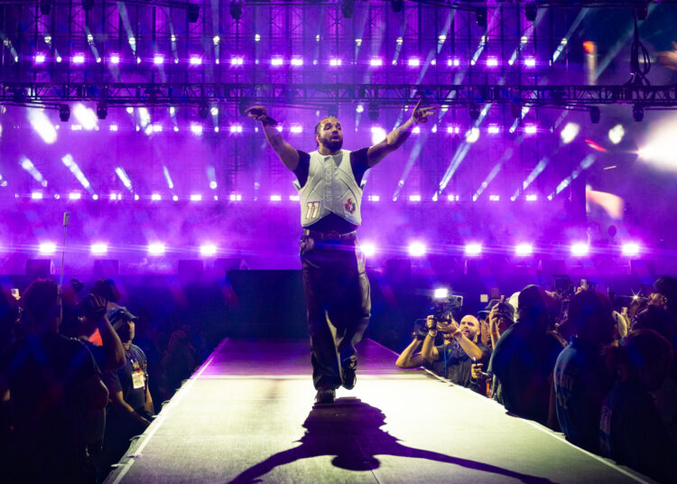 Drake energetically walks down a stage runway with purple lights and a cheering crowd.