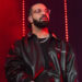 Drake performing on stage, wearing a black leather jacket and clear glasses, with red lighting in the background.