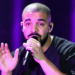 Drake performing on stage, holding a microphone, with a purple-lit background.