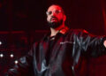 Drake performing on stage, wearing a black leather jacket and glasses, with a red-lit background.