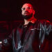 Drake performing on stage, wearing a black leather jacket and glasses, with a red-lit background.