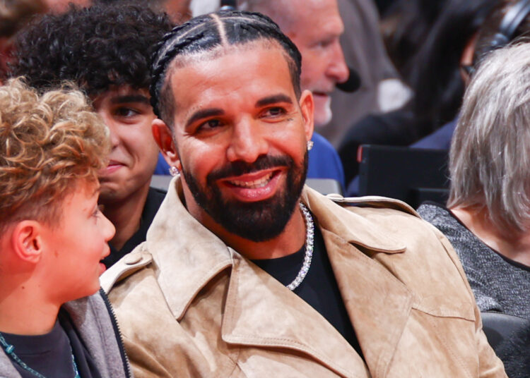 Drake, wearing a tan jacket, smiles while sitting at an event with his son, Adonis, who has curly blond hair.
