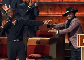 Kevin Hart, in black, holding a microphone, gestures towards Katt Williams in a sparkly jacket and hat on stage at a comedy roast event.