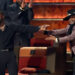 Kevin Hart, in black, holding a microphone, gestures towards Katt Williams in a sparkly jacket and hat on stage at a comedy roast event.