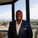 Byron Allen smiling in a dark blue suit in an office with a window view overlooking Los Angeles