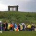 A long queue of people in lots of different coloured raincoats and holding several umbrellas. They are standing in a field with hills in the background. At the top of the hill is the Penshaw Monument which was built in the style of an ancient Greek temple.