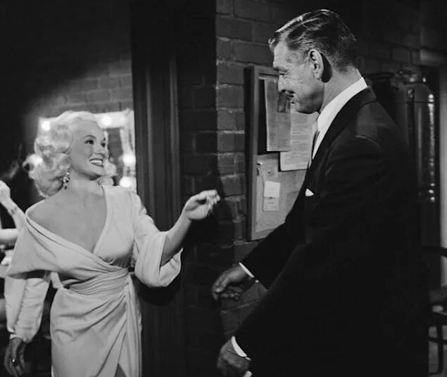 In a monochromatic image, a formally dressed man and woman engage in an intense conversation. The man, dressed in a suit, appear