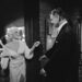In a monochromatic image, a formally dressed man and woman engage in an intense conversation. The man, dressed in a suit, appear