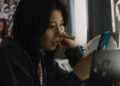 Joan, played by actor Shirley Chen, sits in her room looking at her phone with a clothes pin on her nose in Slanted.