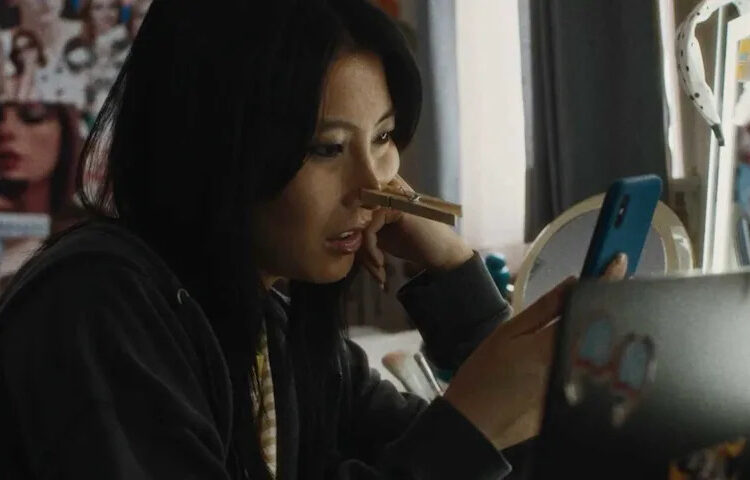 Joan, played by actor Shirley Chen, sits in her room looking at her phone with a clothes pin on her nose in Slanted.