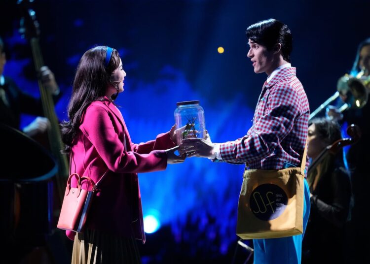 Two people sing to each other on stage while holding a jar