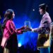 Two people sing to each other on stage while holding a jar