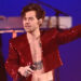 Harry Styles performing on stage, wearing a red sequin jacket, holding a microphone, with tattoos visible on his chest.