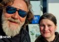 A man with a grey beard and shaggy brown hair. He's wearing mirrored sunglasses and is posing for a selfie with a young girl. She has dark hair and is wearing a black hoodie. They are both smiling.