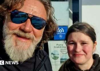 A man with a grey beard and shaggy brown hair. He's wearing mirrored sunglasses and is posing for a selfie with a young girl. She has dark hair and is wearing a black hoodie. They are both smiling.