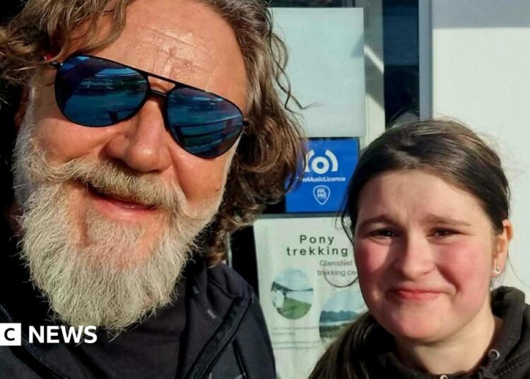 A man with a grey beard and shaggy brown hair. He's wearing mirrored sunglasses and is posing for a selfie with a young girl. She has dark hair and is wearing a black hoodie. They are both smiling.