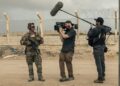 A man holding a camera and a man holding a large mic talk to a service member.