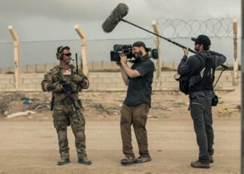 A man holding a camera and a man holding a large mic talk to a service member.