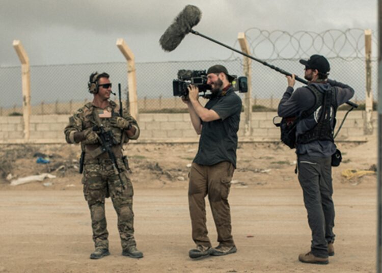 A man holding a camera and a man holding a large mic talk to a service member.