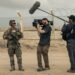 A man holding a camera and a man holding a large mic talk to a service member.