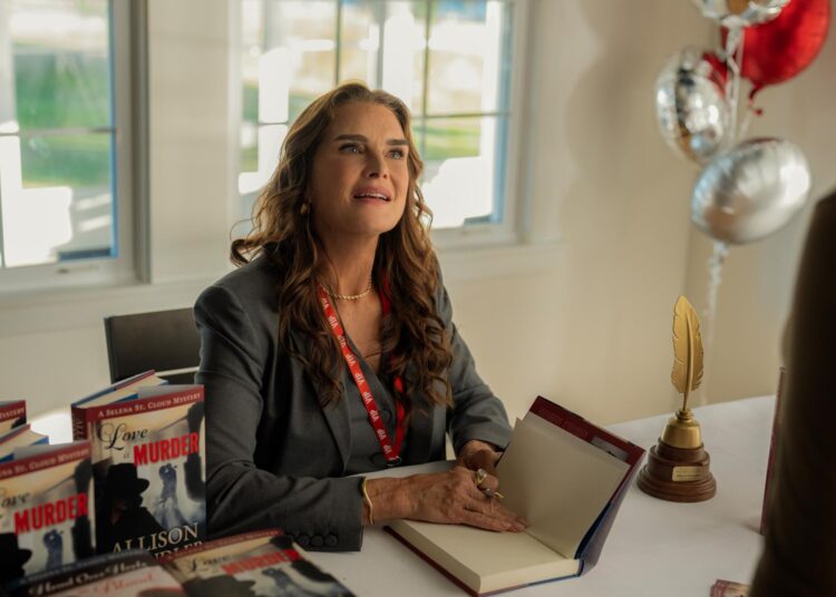 A seated brunet woman in a suit jacket holds a book and gazes up at someone.