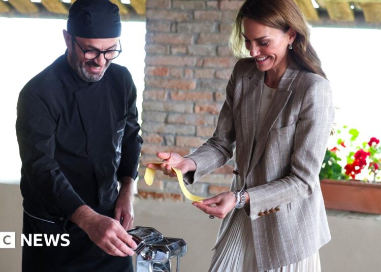 An Italian chef dressed in all black kitchen wear and hat stands to the left of the image next to the Princess of Wales as he helps her make fresh pasta in a kitchen. The Royal, dressed in a grey blazer and skirt is grinning as she holds the pasta in her hands