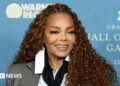 Smiling Janet Jackson stood at a press event against a blue backdrop
