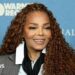 Smiling Janet Jackson stood at a press event against a blue backdrop