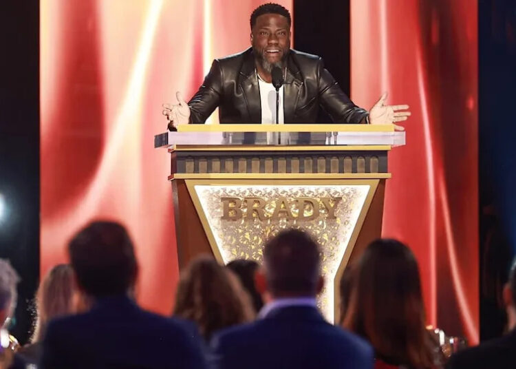 Kevin Hart speaks animatedly while standing at a podium labeled "BRADY" in front of an audience during an event. He wears a sharp black leather jacket