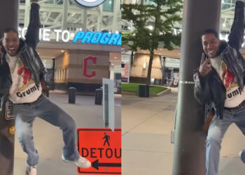 Kid Cudi playfully hangs from a pole in front of a stadium entrance with "Welcome to Progressive" signage, wearing a leather jacket and jeans.