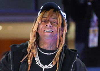 Lil Wayne smiling, wearing a black cap, dreadlocks, two diamond chains, and a grill.