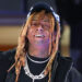 Lil Wayne smiling, wearing a black cap, dreadlocks, two diamond chains, and a grill.