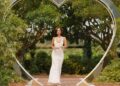 An individual dressed in a white gown stands in the midst of a heart-shaped structure, surrounded by lush greenery and a backdrop of trees.