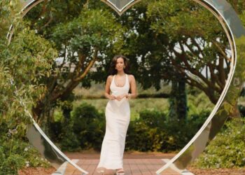 An individual dressed in a white gown stands in the midst of a heart-shaped structure, surrounded by lush greenery and a backdrop of trees.