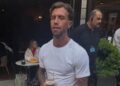 A man with slicked back hair and white t shirt holds a half drunk pint