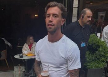 A man with slicked back hair and white t shirt holds a half drunk pint