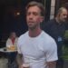 A man with slicked back hair and white t shirt holds a half drunk pint