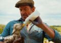 Hugh Jackman playing a shepherd. He's wearing a brown flat cap with a blue jacket with rolled up sleeves. Underneath he has a white shirt. He's looking down at and cradling a muddy lamb in his right arm while feeding it with a plastic milk bottle.