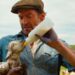Hugh Jackman playing a shepherd. He's wearing a brown flat cap with a blue jacket with rolled up sleeves. Underneath he has a white shirt. He's looking down at and cradling a muddy lamb in his right arm while feeding it with a plastic milk bottle.