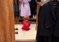 A woman in a lavender coat stands in front of a mirror, while a child in a red outfit sits on the floor.